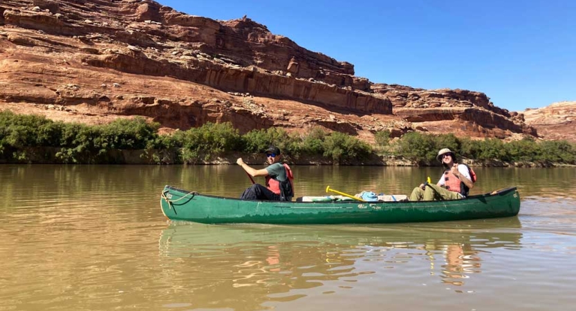 Two people wearing life jackets paddle a canoe on calm water. On the other side of the river are tall red canyon walls. 